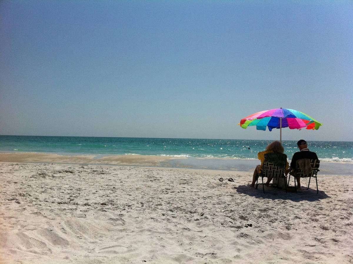 Side-by-side comparison showing UV index readings under a standard beach umbrella (UVI 4.2) versus a UPF 50+ umbrella (UVI 0.8), with reflected UV rays illustrated bouncing off sand and water